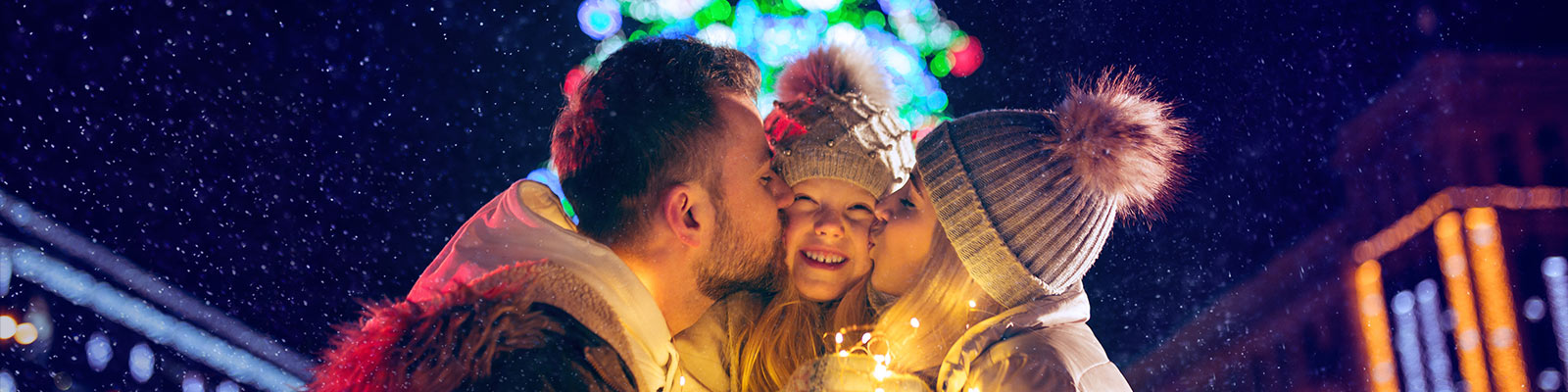 Un padre y una madre dan un beso a su hija. De fondo un arbol de navidad iluminado.